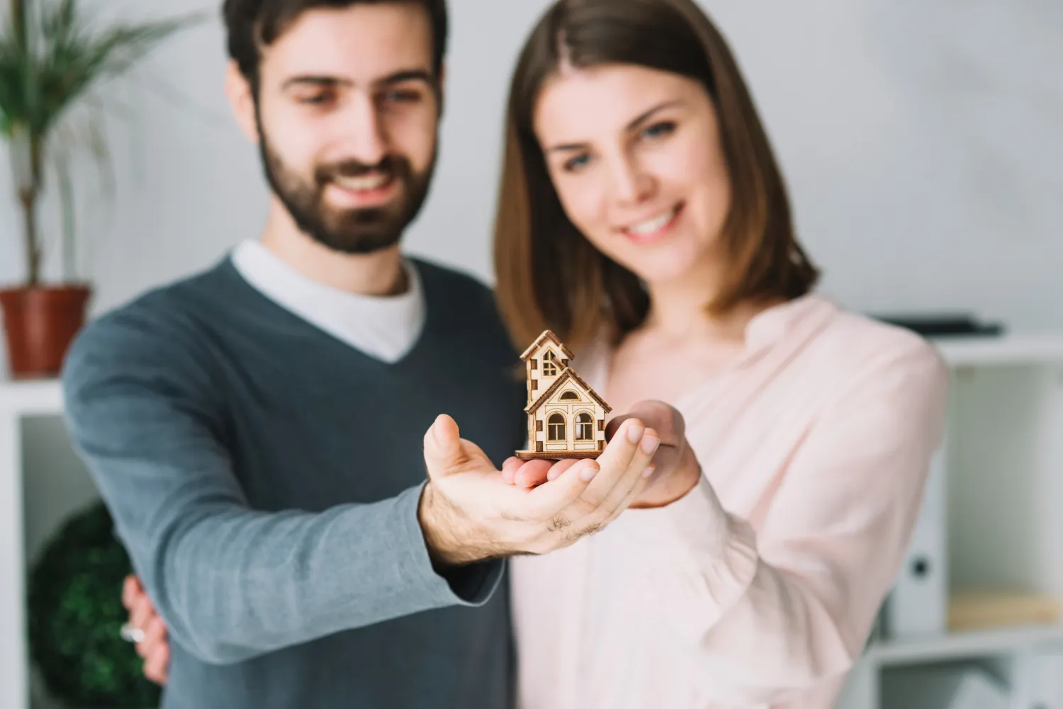 Pareja sonriente sosteniendo una casa de juguete en sus manos, simbolizando la compra de una nueva vivienda. Fondo con elementos decorativos y plantas.