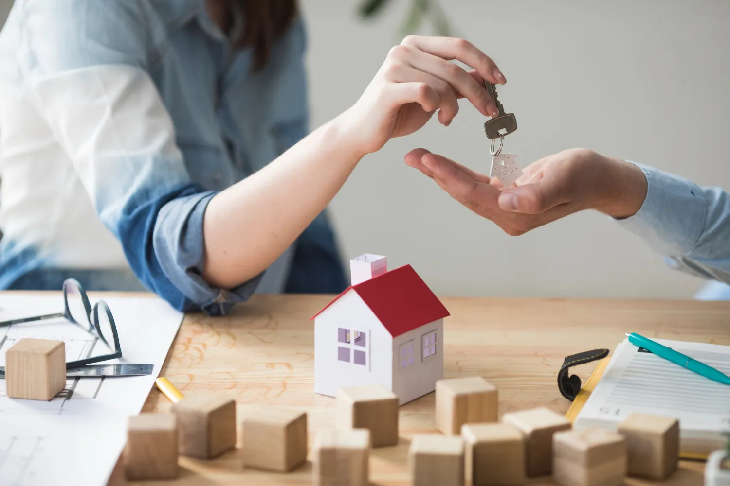 Una mano entrega una llave a otra persona sobre una mesa, con una casa de papel en miniatura y bloques de madera alrededor, simbolizando la compra de una vivienda.