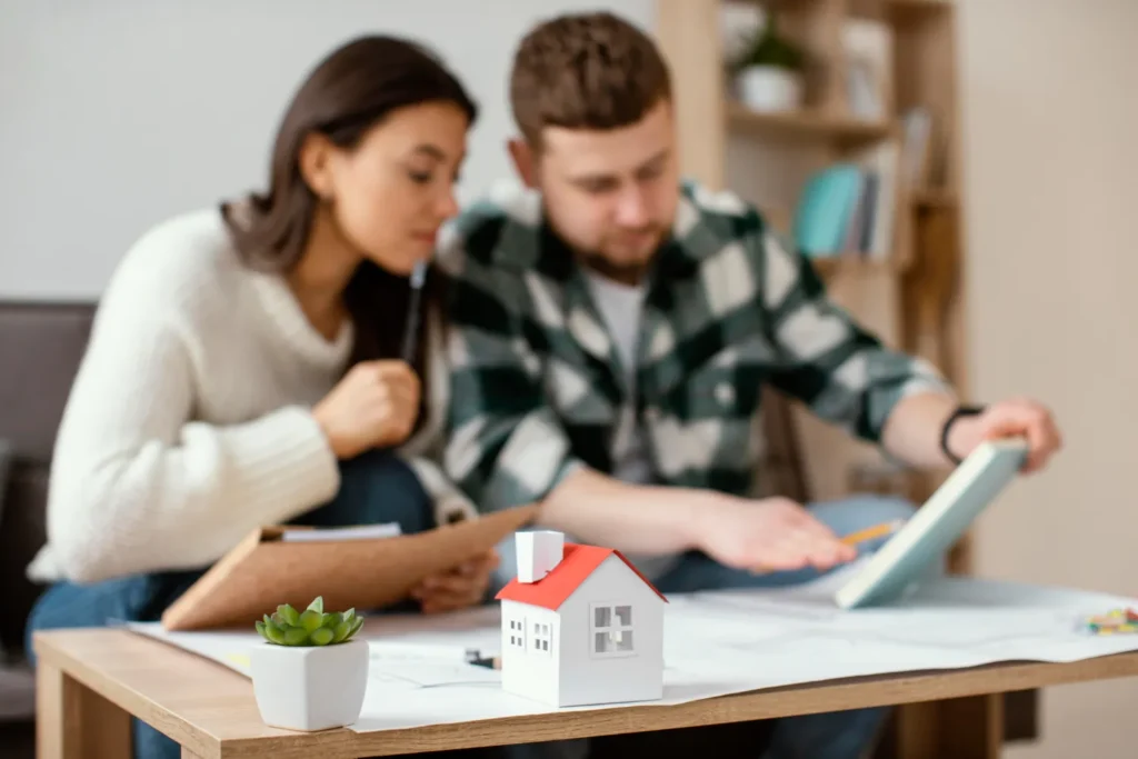 Pareja revisando planos de construcción en una mesa, con un modelo de casa en miniatura y una planta suculenta en primer plano.